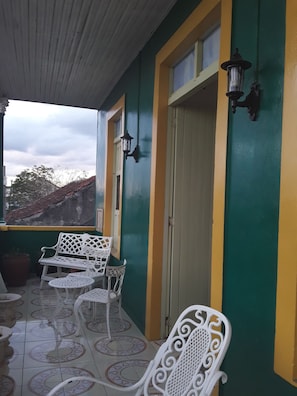 A covered patio area with white metal furniture, including benches and tables, against a green and yellow wall. - Casa El Mirador - Yliana (Baracoa)