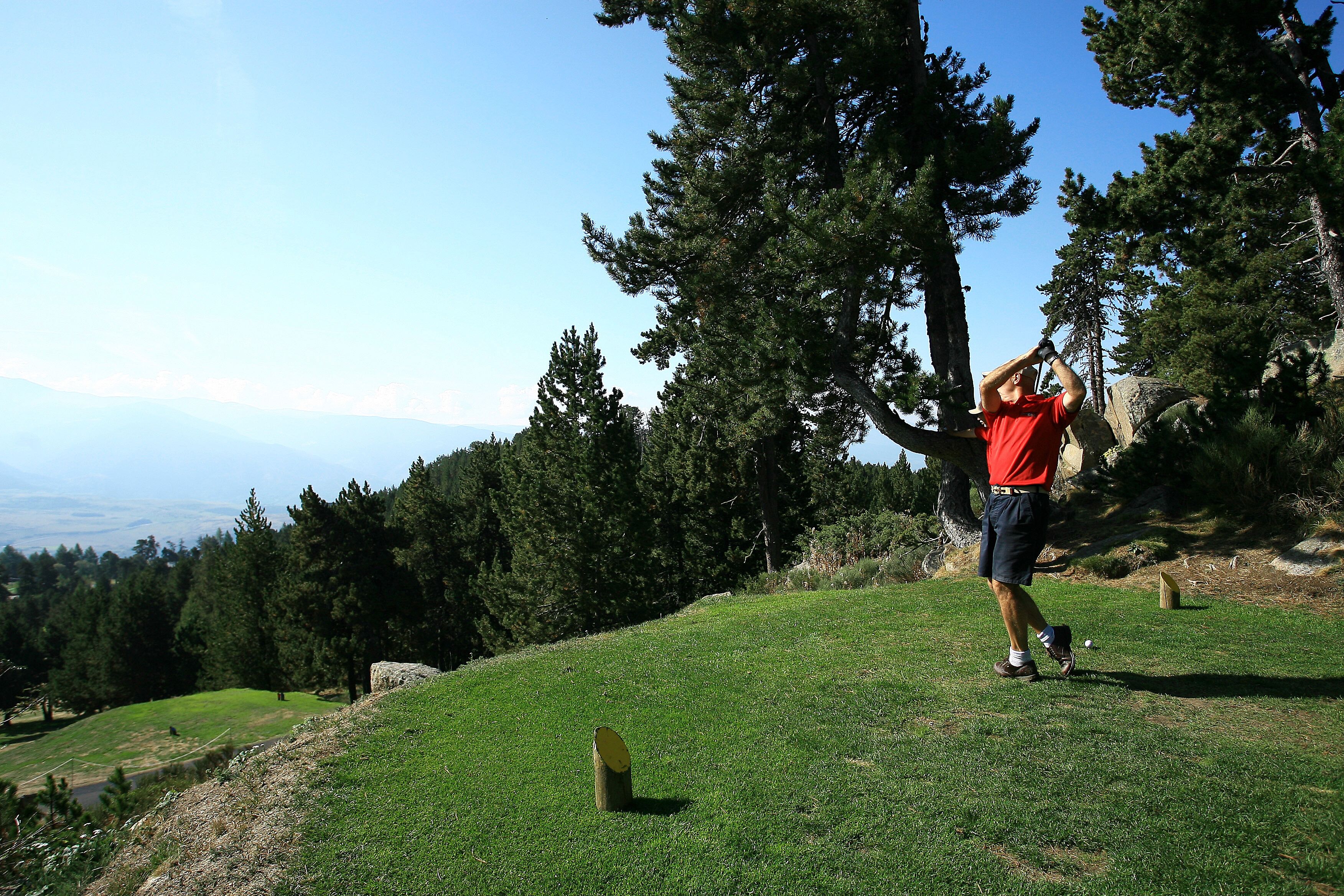 Uma pessoa jogando golfe numa colina gramada, com árvores e um céu limpo ao fundo.
