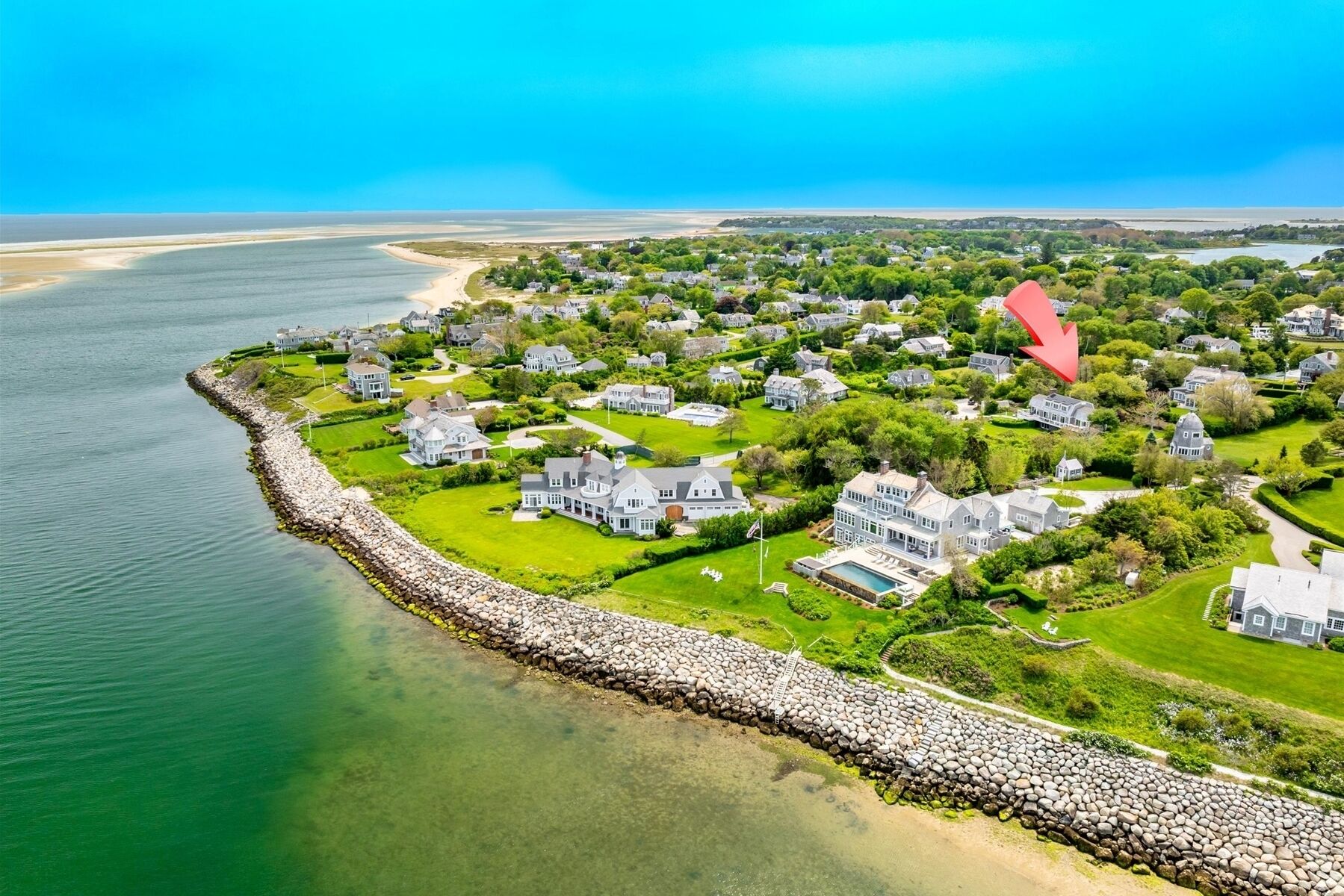 An aerial view of a coastal residential area with houses, a stone seawall, and a clear view of the ocean.