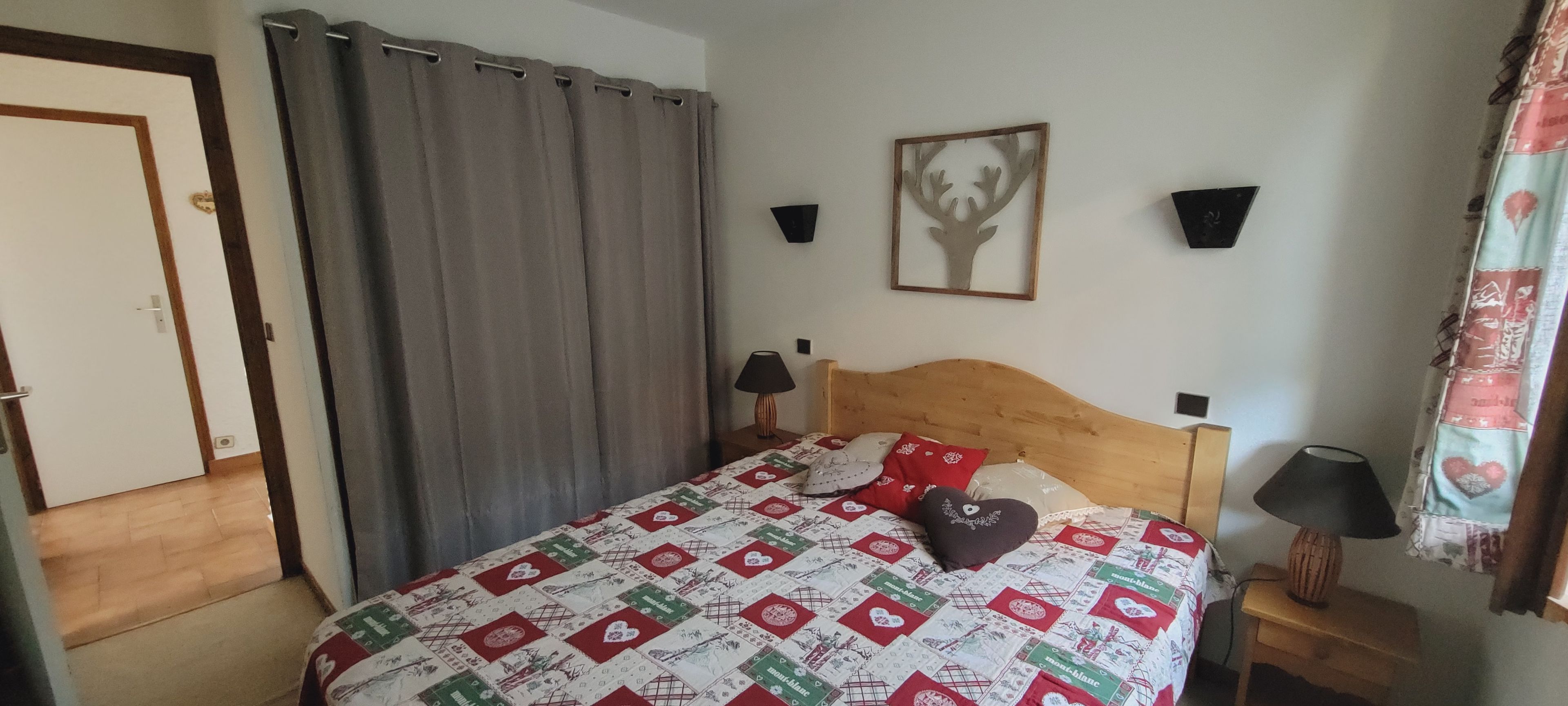 A bedroom with a bed, a wooden headboard, a nightstand with a lamp, a wall-mounted shelf, and a door.