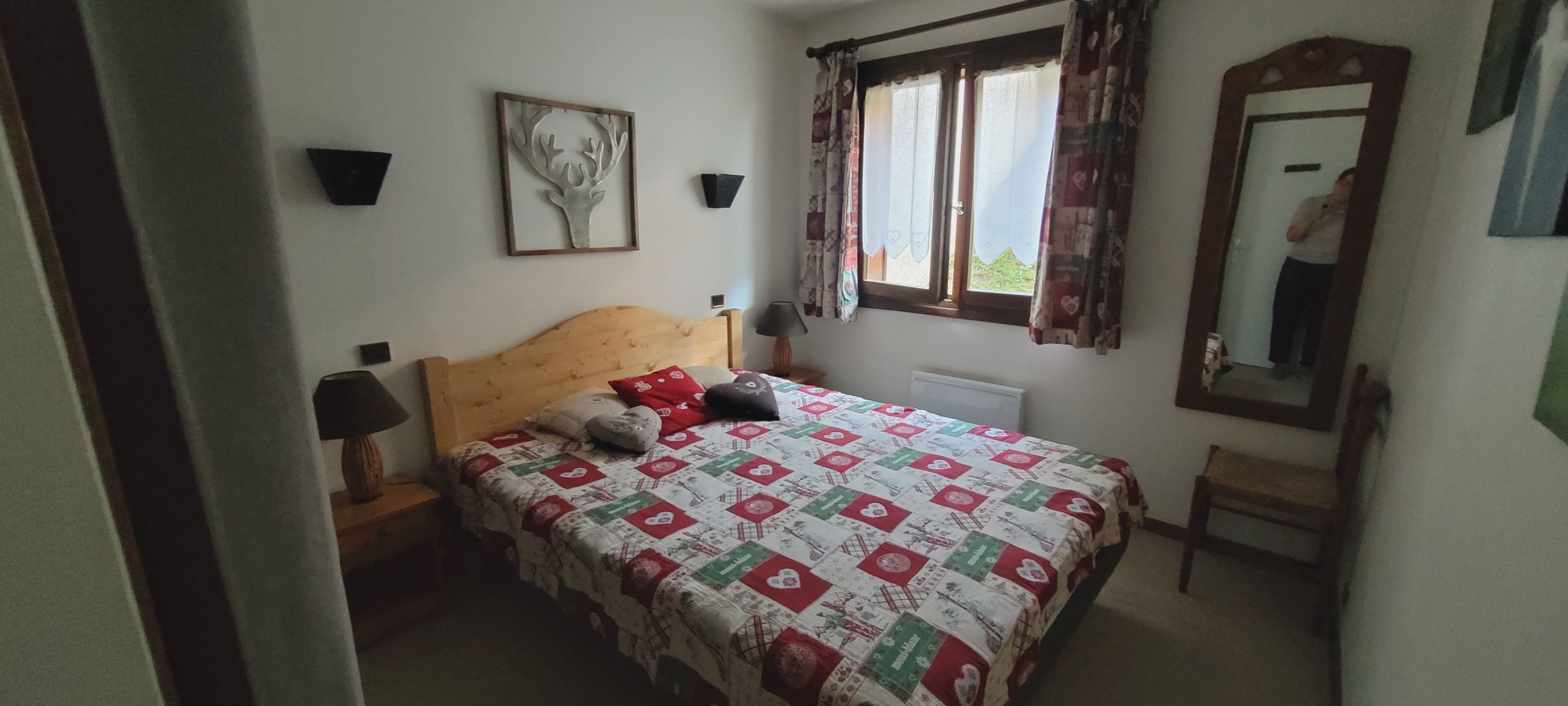 A bedroom with a bed, a nightstand, a lamp, a window with curtains, a mirror, and a wall-mounted picture.