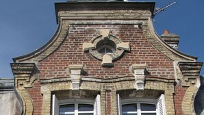 Ein Gebäude mit einer symmetrischen Fassade, ein zentrales Fenster mit Fensterläden, flankiert von zwei kleineren Fenstern mit Fensterläden, und ein dekoratives Steinwappen darüber.