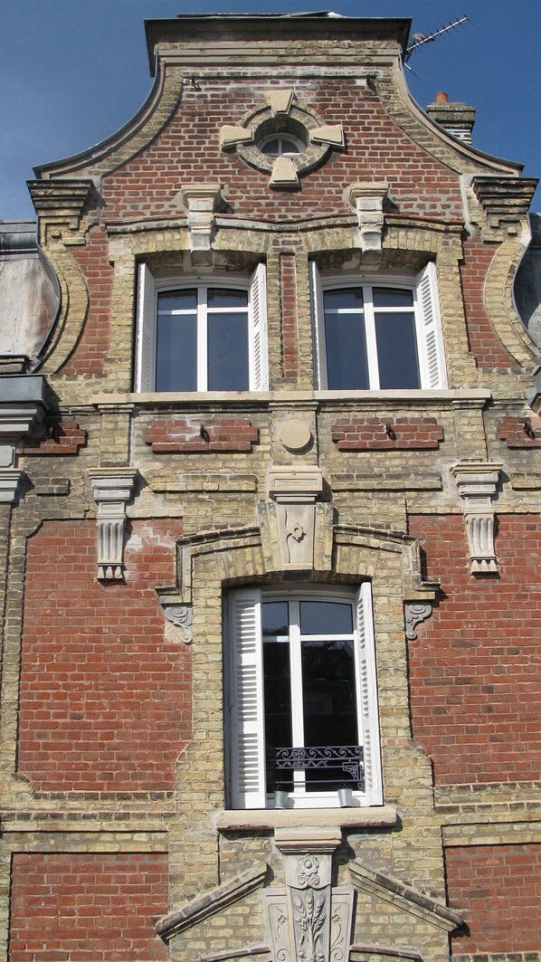 A building with a symmetrical facade, featuring a central window with shutters, flanked by two smaller windows with shutters, and a decorative stone crest above.
