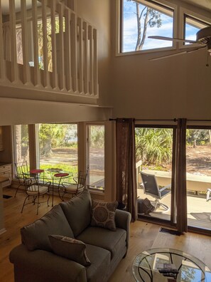 A spacious living room with a high ceiling, large windows, a dining area with a table and chairs, and a view of the outdoors.