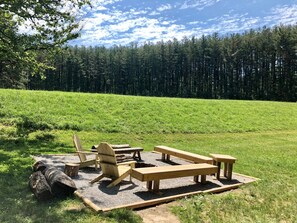 Un espace pique-nique aménagé avec des bancs et des chaises, situé dans un champ herbeux, avec une forêt en arrière-plan.