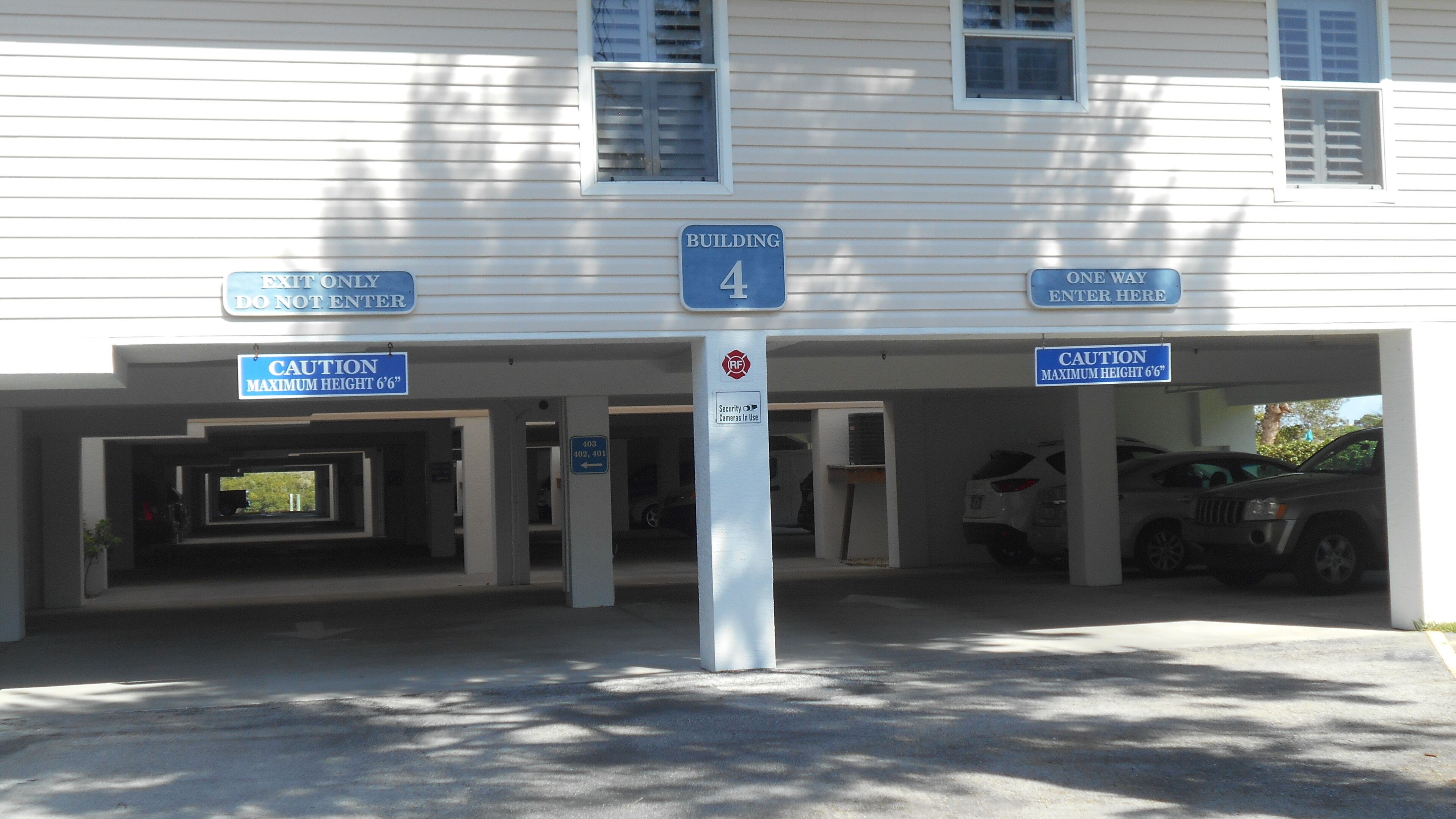 Entrada de un edificio con carteles que indican 'Edificio 4', 'Precaución Altura Máxima 6 pies 6 pulgadas', 'Cámaras de Seguridad en Uso' y 'Solo Ida, Entre Aquí'.