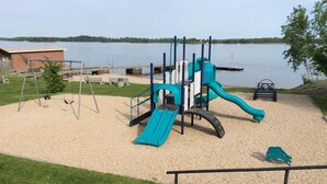 Un parque infantil con tobogán y columpios, rodeado de una zona de arena y un lago al fondo.