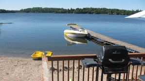 Una terraza de madera con una parrilla, un bote amarillo y un muelle que se adentra en un lago tranquilo.