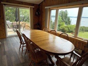 Une salle à manger en bois avec une grande table et des chaises, de grandes fenêtres donnant sur un lac et des arbres, et une porte-fenêtre coulissante qui mène à un patio extérieur.