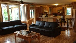 Un salón acogedor con un sofá de cuero negro, una mesa de centro de vidrio y suelo de madera. Se aprecia una zona de cocina al fondo, con armarios de madera y un microondas.