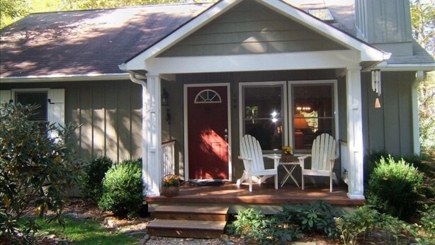 Une maison avec un porche à l’avant, deux fauteuils Adirondack blancs et une porte rouge.