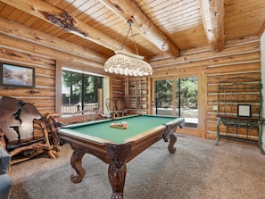 Interno di una baita in legno con tavolo da biliardo, sedie in legno, un lampadario e un'ampia finestra.