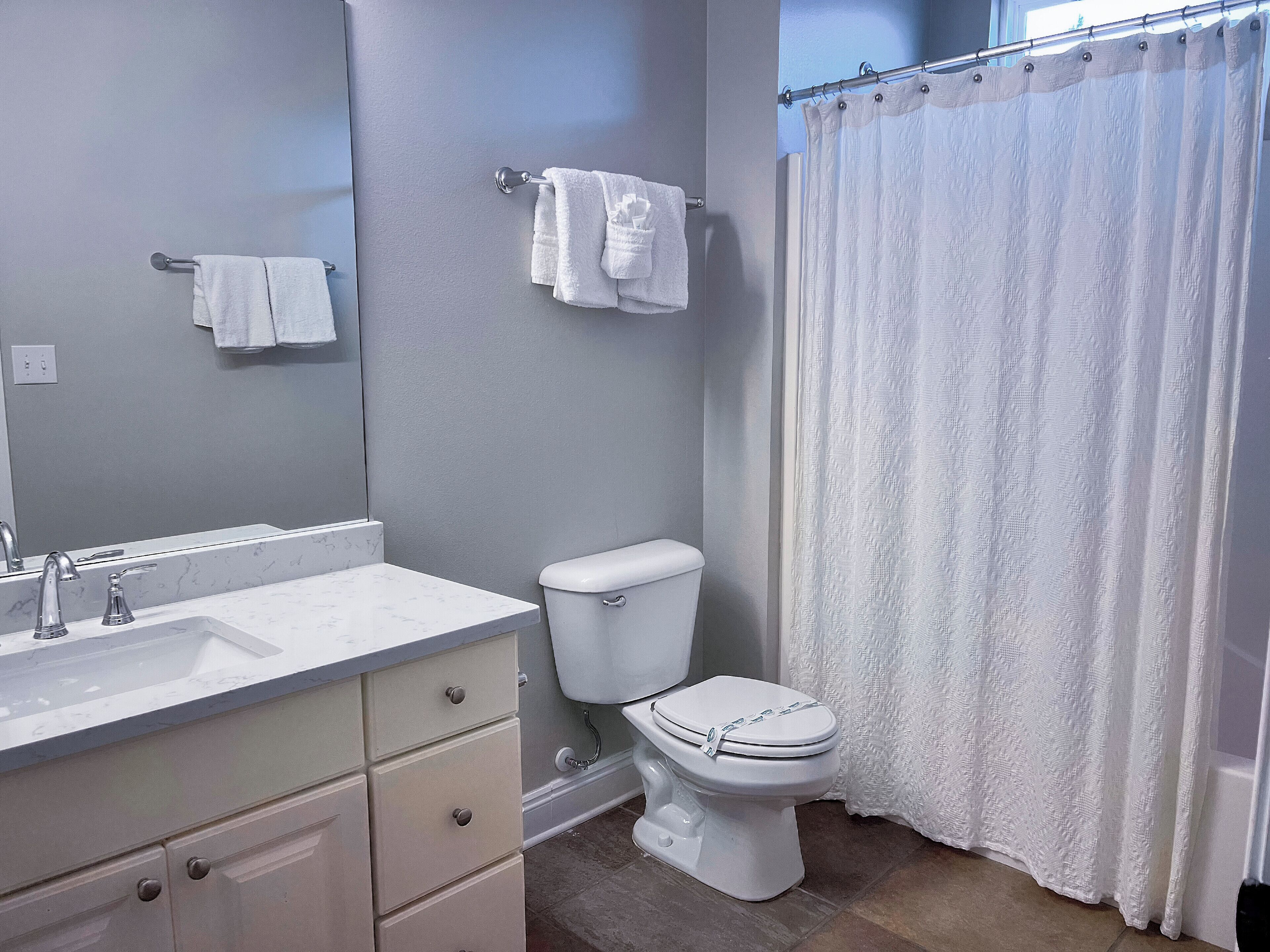 Ein Badezimmer mit weißem Waschbecken, Spiegel und Toilette. Handtücher hängen an einem Ablagegestell und ein Duschvorhang ist angebracht.