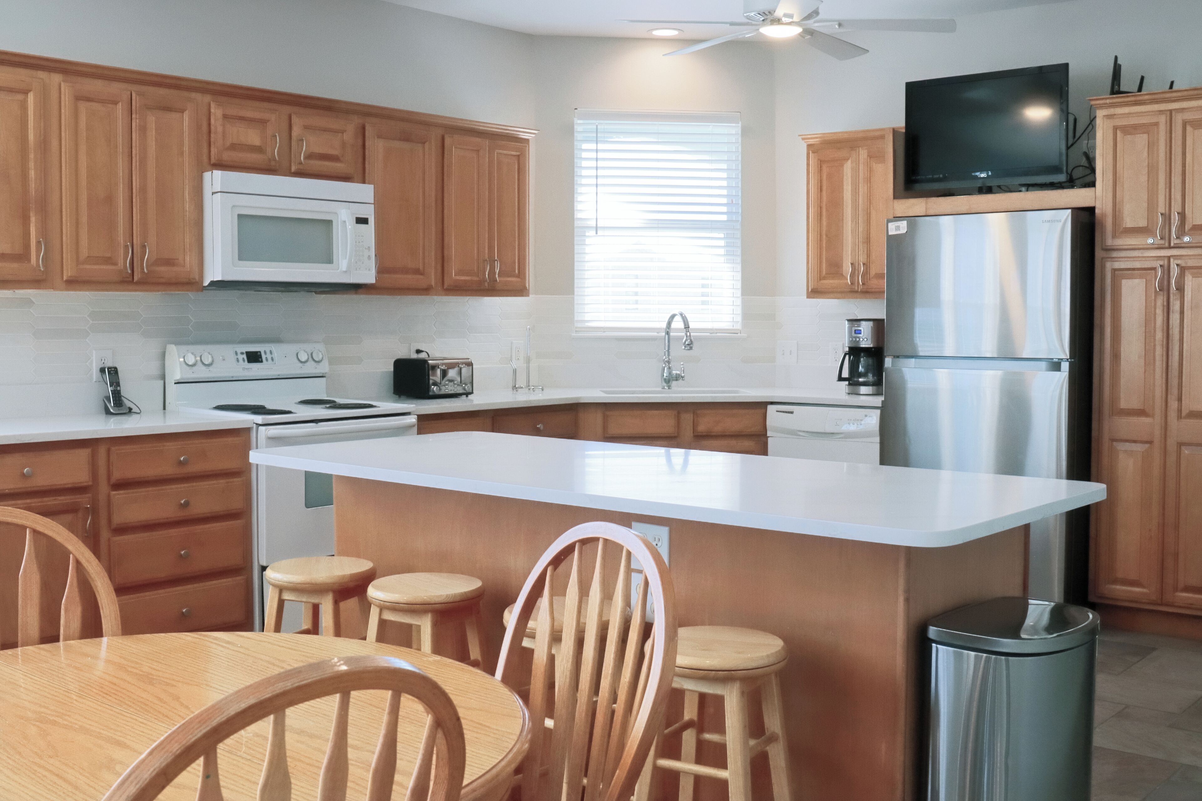 A modern kitchen with wooden cabinets, a white countertop, a stainless steel refrigerator, a microwave, a stove, and a dining area with wooden chairs and stools.