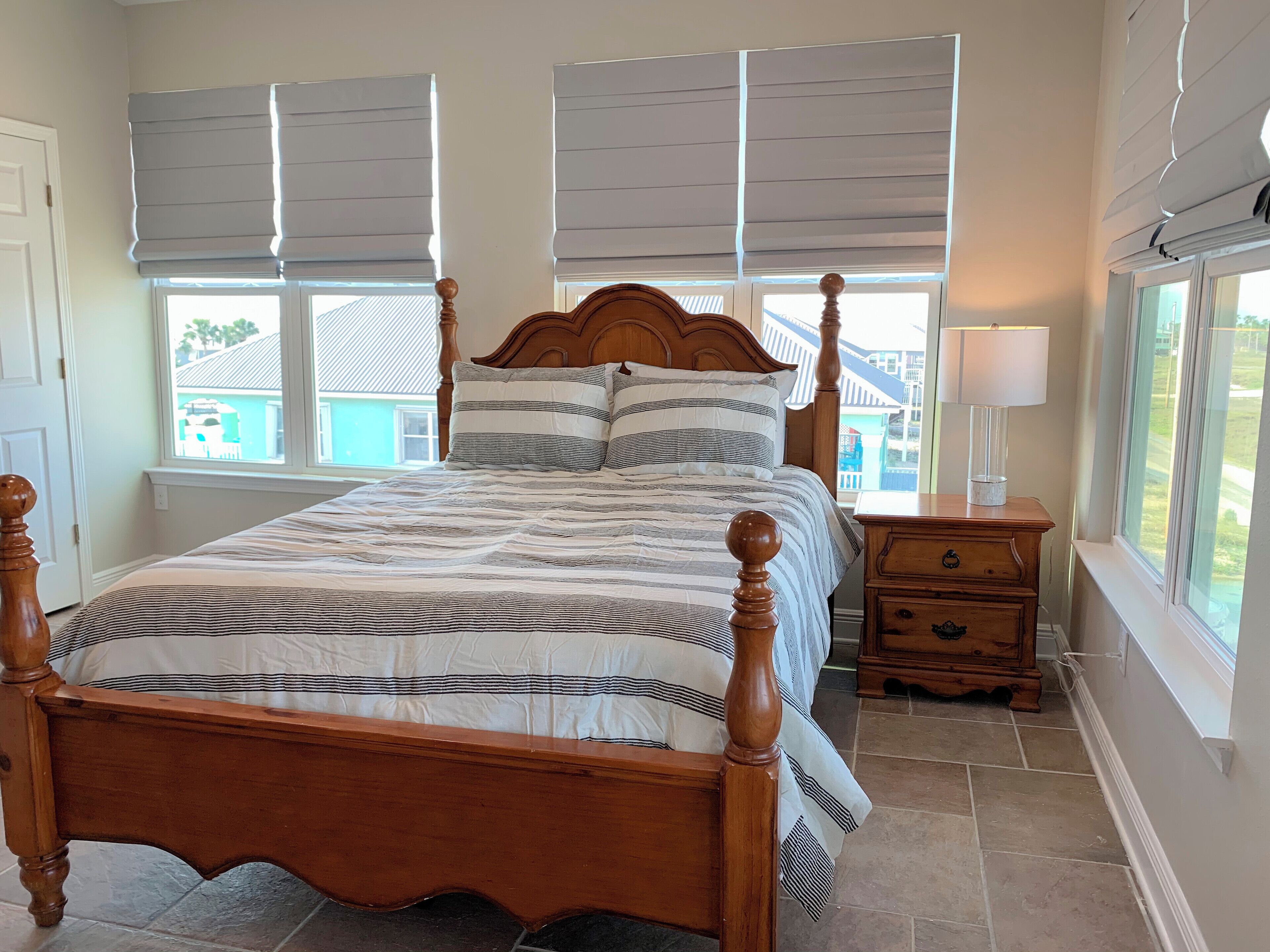 Un dormitorio con una cama de madera, ropa de cama a rayas, una mesita de noche y ventanas amplias con persianas.