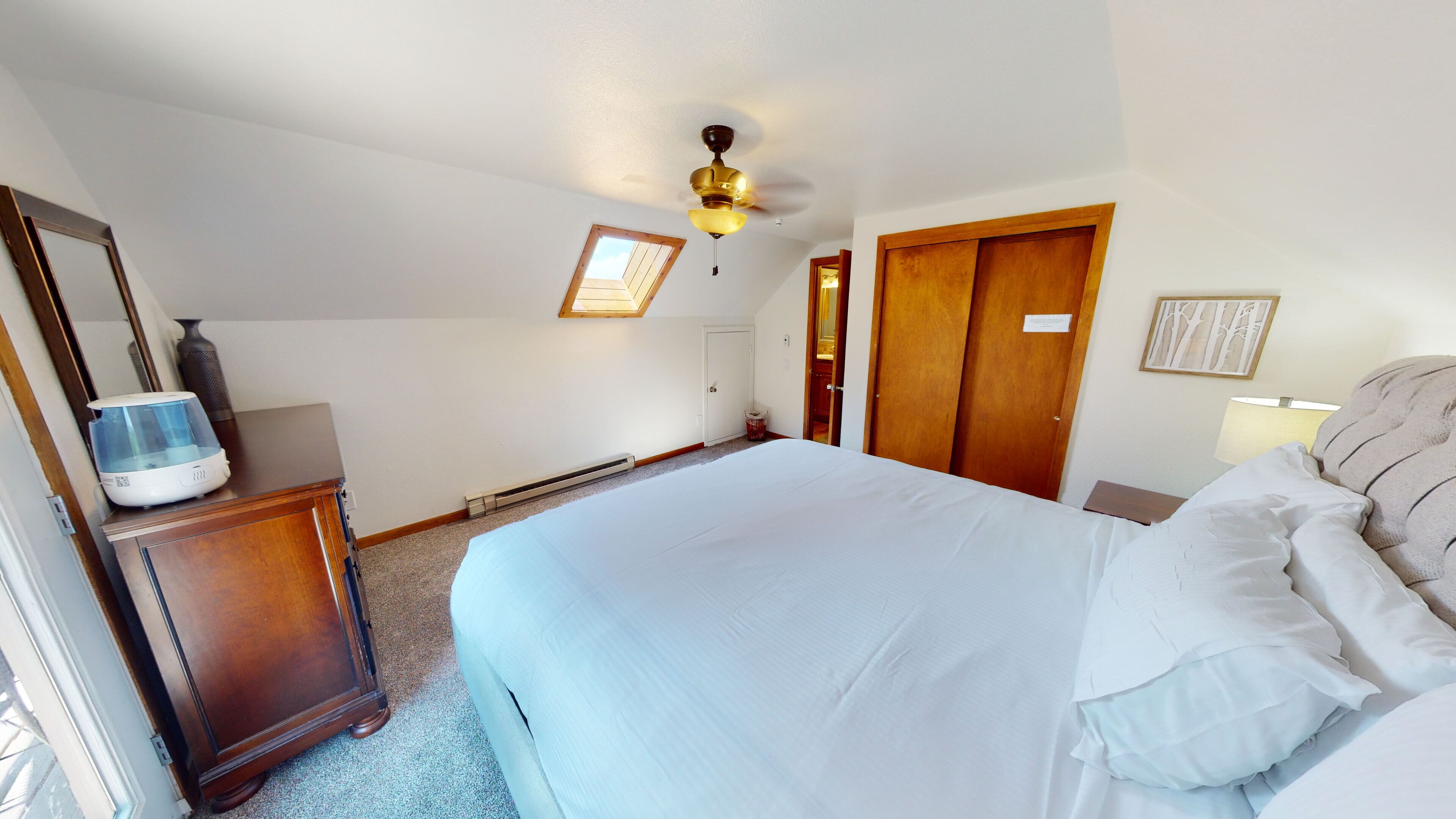 A bedroom with a bed, wooden wardrobe, a desk with a water dispenser, a ceiling fan, and a small window.