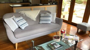Una sala de estar moderna con un sofá gris, cojines estampados, una mesa de centro de vidrio con revistas y vistas al exterior a través de amplios ventanales.