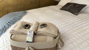 Una cama bien hecha con un edredón blanco texturizado, una manta gris y una cesta tejida con toallas dobladas y una botella de champú.
