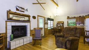 A cozy living room with a fireplace, wooden floors, and a dining area with a kitchen in the background. - WIFI, HOT TUB, ~A/C~, SMART TV IN BD. FIRE PIT BY THE CREEK, 2 MIN TO MAGGIE (Maggie Valley)