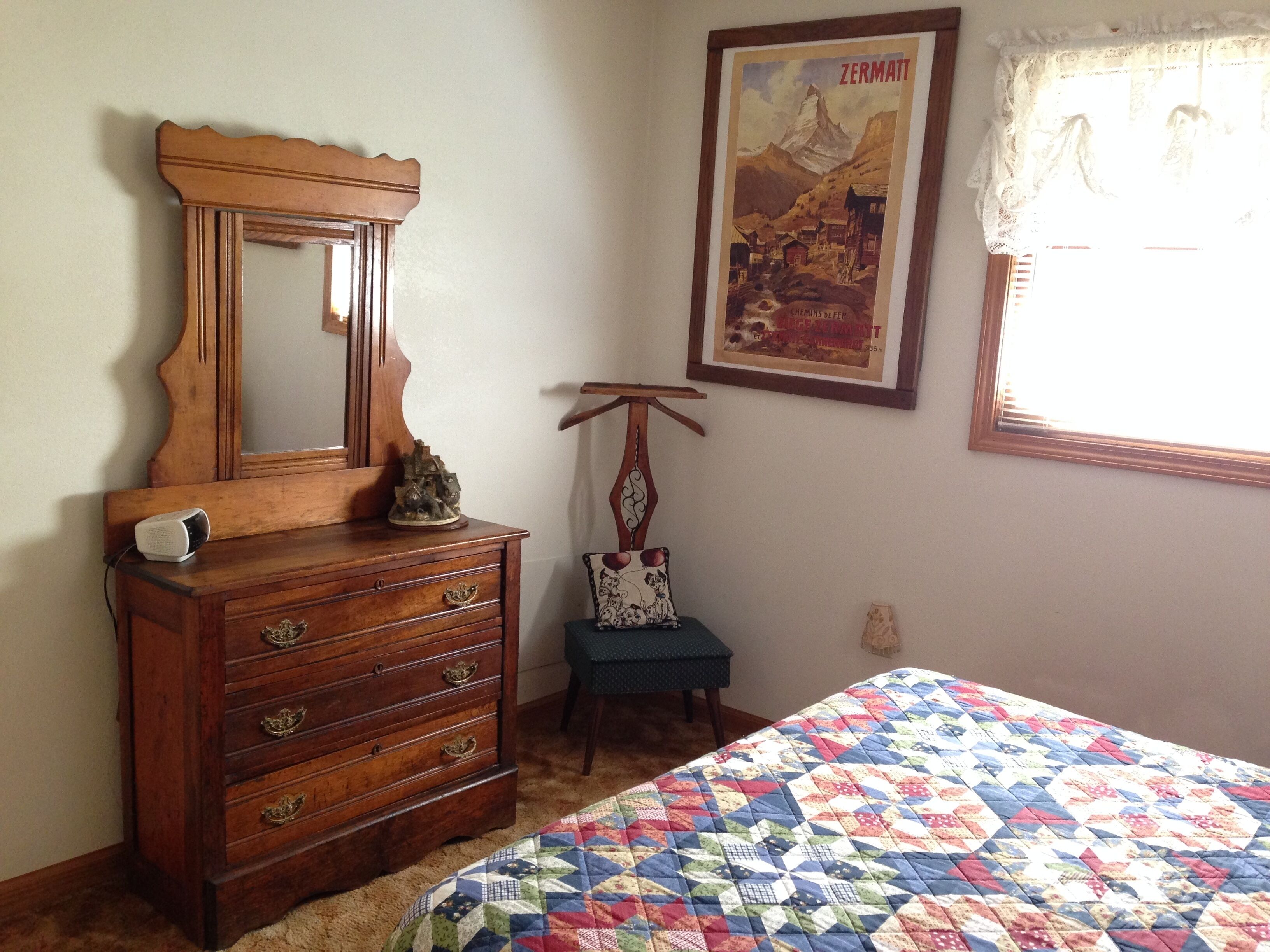 Une chambre avec une commode en bois, un lit recouvert d’une courtepointe aux couleurs vives, une affiche accrochée au mur et une fenêtre ornée de rideaux en dentelle.