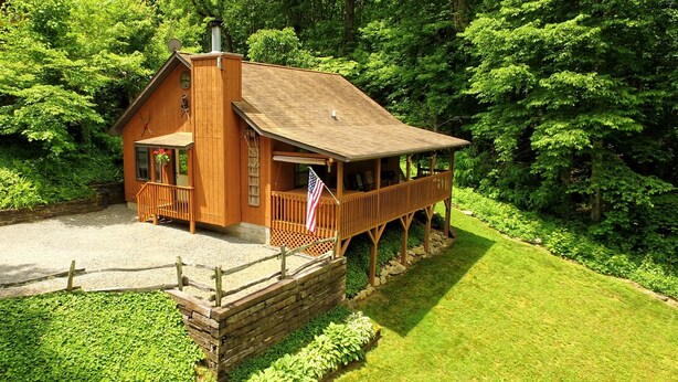 A wooden cabin with a porch and a flag, surrounded by lush greenery. - WIFI, HOT TUB, ~A/C~, SMART TV IN BD. FIRE PIT BY THE CREEK, 2 MIN TO MAGGIE (Maggie Valley)