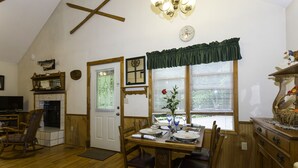 A dining area with a wooden table set for four, a wooden chair, a clock on the wall, and a window with blinds. - WIFI, HOT TUB, ~A/C~, SMART TV IN BD. FIRE PIT BY THE CREEK, 2 MIN TO MAGGIE (Maggie Valley)