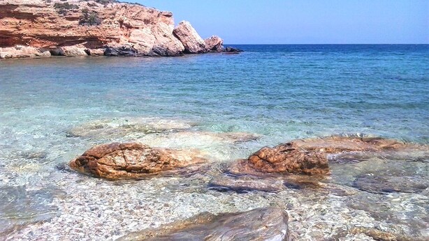A coastal scene with clear shallow waters revealing rocks and a rocky cliff in the background. - Villa Mella, Paros: Idyllic waterfront Villa that combines privacy & relaxation. (Paros)