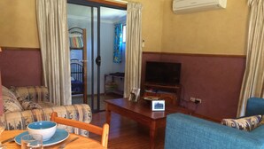 Una sala de estar con un sofá, una mesa de comedor con cuencos azules y copas de vino, un televisor sobre un mueble de madera y una puerta corrediza de vidrio que da a un balcón.