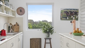 Une cuisine avec des meubles blancs, une fenêtre donnant sur des arbres, une horloge et un tableau représentant des chevaux.
