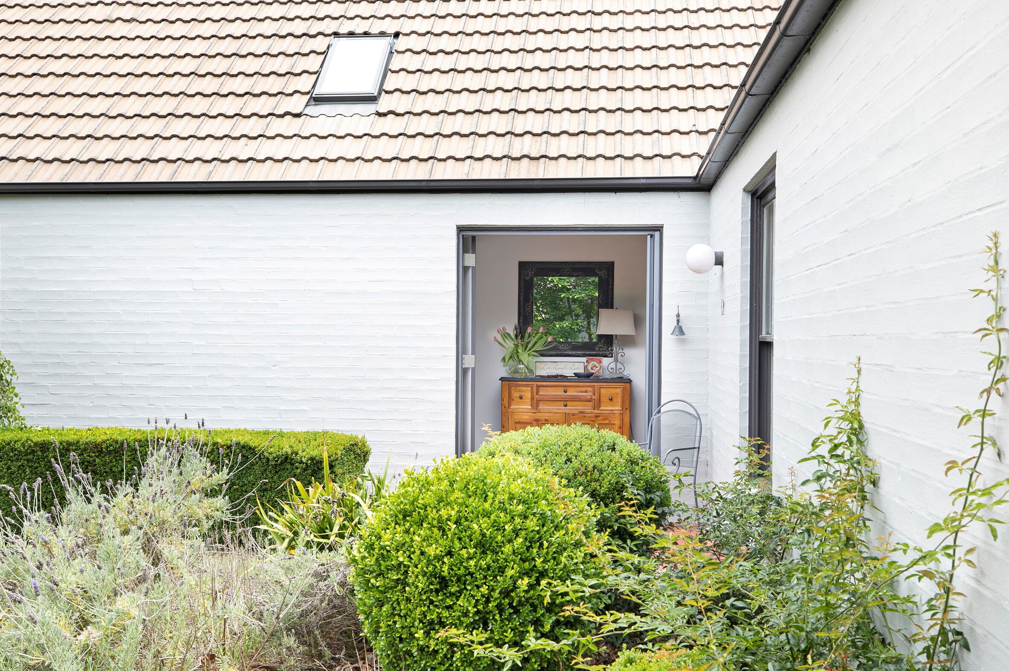 A white building with a tiled roof, a wooden cabinet near the entrance, and various green shrubs and plants around.