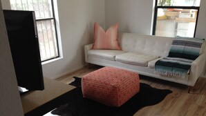 Un salon avec un canapé blanc, un pouf rouge à motifs, un tapis en cuir noir et un téléviseur posé sur un meuble.