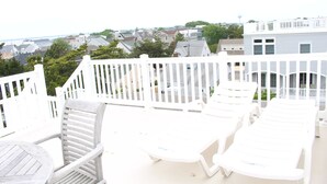 A balcony with white plastic chairs and a wooden chair, overlooking a residential area with houses and trees.