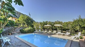 Piscina con sillas de descanso y sombrillas, rodeada de follaje y una escalera de piedra.