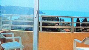 A balcony with a view of the sea, a table with a hat and a floral tablecloth, and two white chairs.