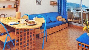 A room with a blue sofa, a dining table with chairs, and a view of the sea through a window.