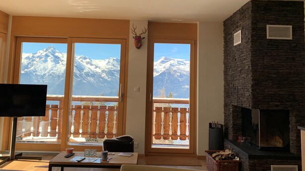 A living room with a fireplace, a television, and a view of snow-capped mountains through large windows.
