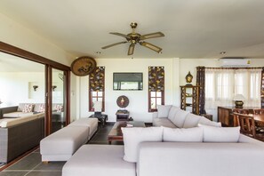 A spacious living room with a large sofa, a coffee table, and a ceiling fan. There are two windows with curtains and a wall-mounted TV.