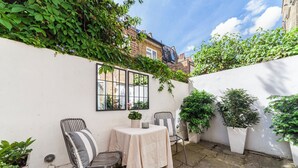 A small outdoor patio with a table and two chairs, surrounded by potted plants and a white wall. - Cool 1 bed in Munster village fulham. (London)