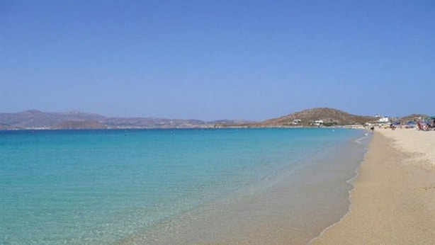 Uma praia com águas turquesa cristalinas, faixa de areia e colinas ao longe.