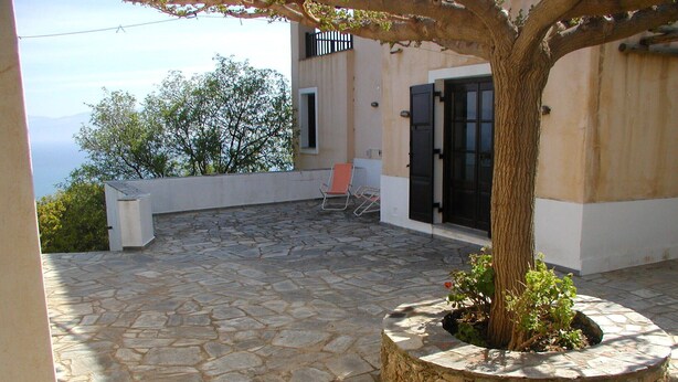 A stone patio with a tree planter, a white building with a black door, and a red chair. - Country villa with amazing view, close to important sites and beaches. (Korinthia)