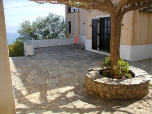 Une terrasse en pierre avec un bac à plantes, un bâtiment blanc avec une porte noire et une chaise rouge.