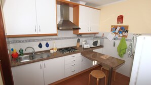 Een praktische keuken met witte kasten, een roestvrijstalen afzuigkap, een spoelbak, een fornuis, een magnetron en een koelkast. Er staan twee krukken bij en op de muur hangt een prikbord met notities en foto’s.