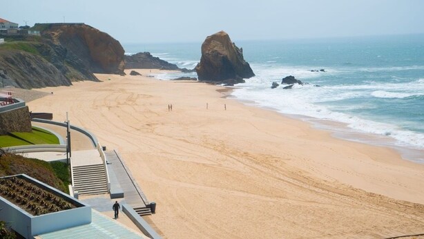 Une plage côtière avec un promontoire rocheux, une étendue de sable et un bâtiment doté d’un toit incurvé et d’un escalier menant à la plage.
