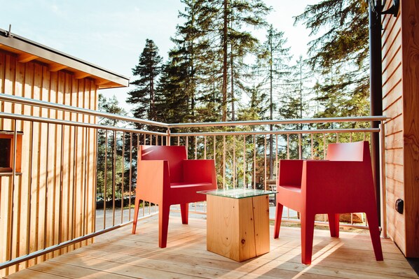 A wooden deck with two red chairs and a glass-top table, surrounded by tall trees.