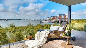 Een terras met een rieten bank, een bijzettafeltje met een vaas en appels, en uitzicht op een jachthaven met boten en gebouwen aan de overkant van het water.