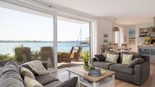 Een ruime woonkamer met een groot raam met uitzicht op een waterpartij en een brug, ingericht met een grijze bank, een houten salontafel en rieten stoelen.