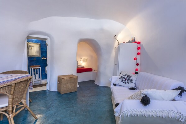 A room with a white wall, a bed, a white sofa, a wicker table, and a small stool.