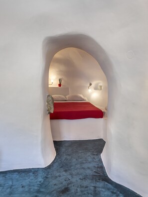 A bedroom with a bed under a white arched ceiling, a red bedspread, and a small red pot on the nightstand.