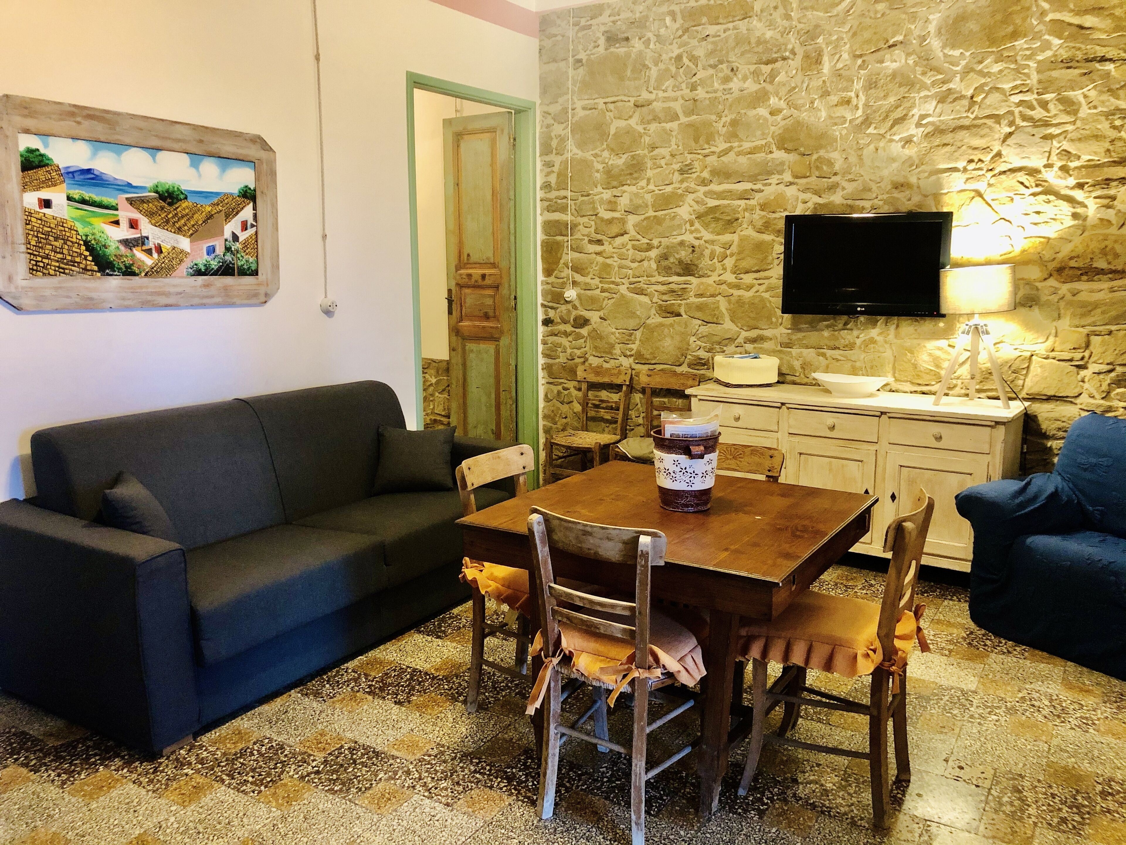 A cozy living area with a stone wall, a wooden dining table, a sofa, and a television mounted on the wall.