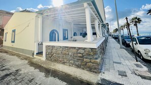 A coastal building with a stone foundation and a white facade, featuring a covered outdoor seating area and a view of the sea.